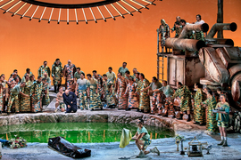 Against an orange background, a crowd of actors wearing makeup and similar camouflage outfits surrounds a circular green pool. A bald man in center is in pain and everyone is turned toward him. A giant rusty machine is on the right.