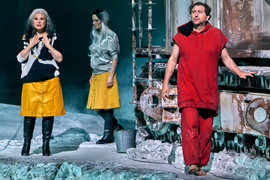 On stage, there are grey rocks and a large rusty machine. A barefoot man dressed in red, and 2 people in yellow skirts and grey hair are on stage, and all seem forlorn.